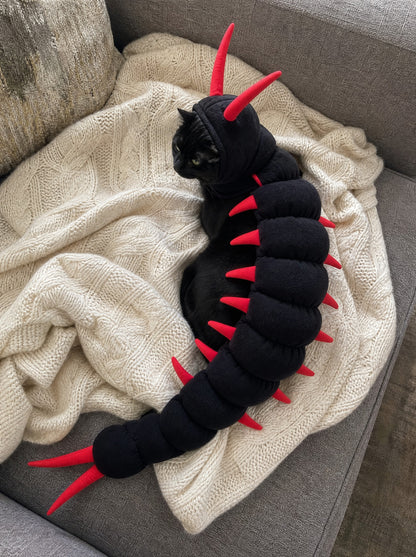 A black cat in the Scuttlecat costume curled up on a knitted throw